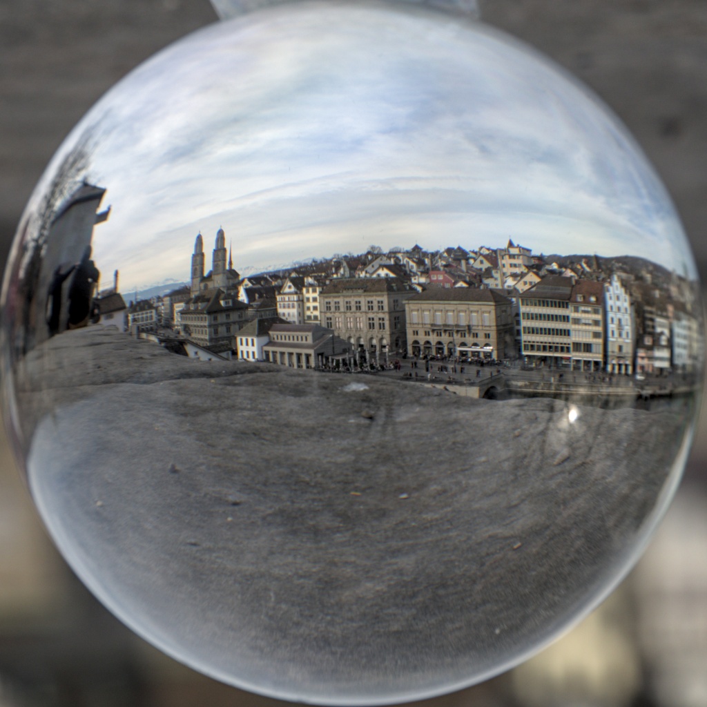 The center of Zürich, seen in a crystal ball. The crystal ball takes all the square picture's area, and shows, with deformed optics, the two towers of Grossmünster, the Alps very faintly in the background, and a bunch of grey/brown buildings.